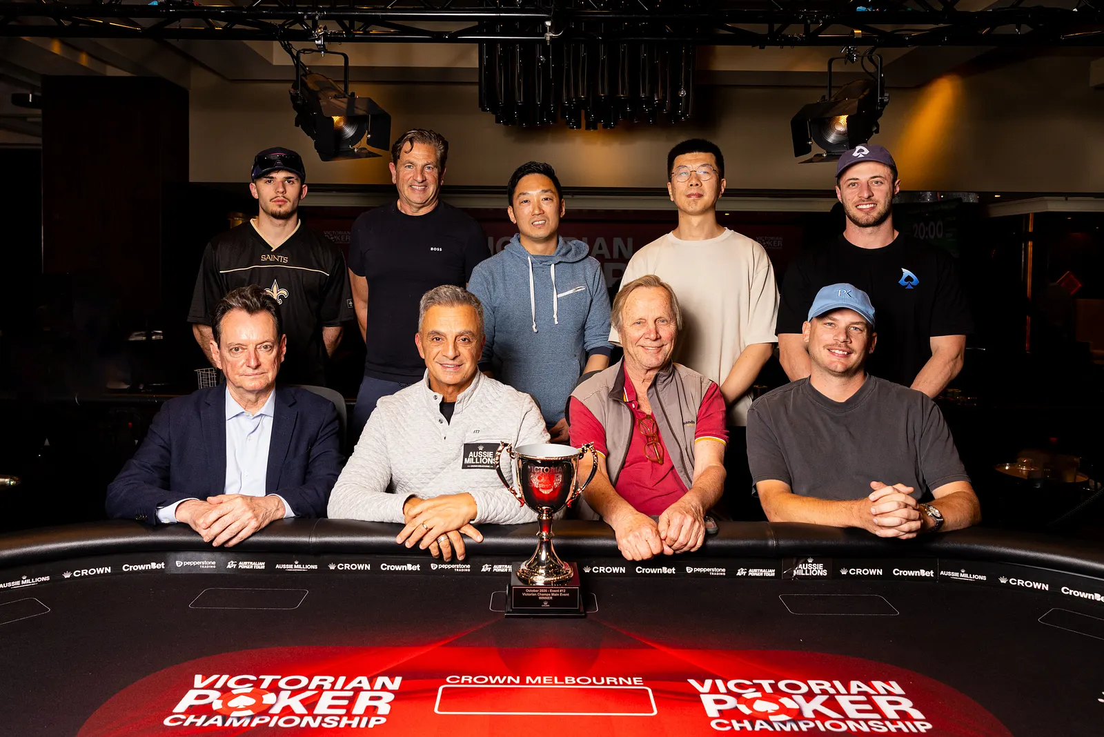 Nove jogadores finalistas do Victorian Poker Championship 2026 posam atrás da mesa de poker com o troféu no centro.